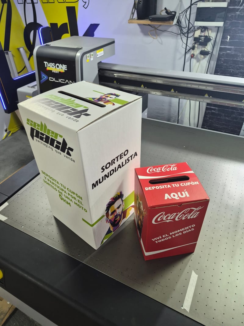 Dos urnas de cartón en la planta de Sellerpack: urna blanca del Sorteo Mundialista con branding Sellerpack y urna roja de Coca-Cola.